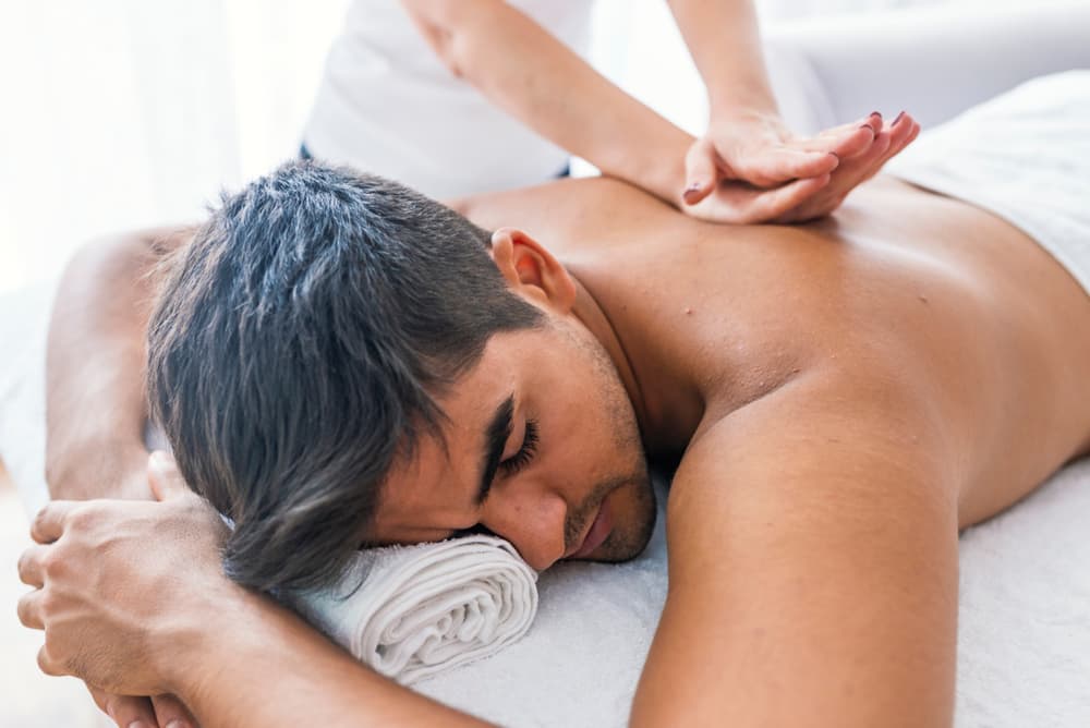 Man lying face down receiving a relaxing full body massage, eyes closed, with natural daylight in a calm and professional treatment room.