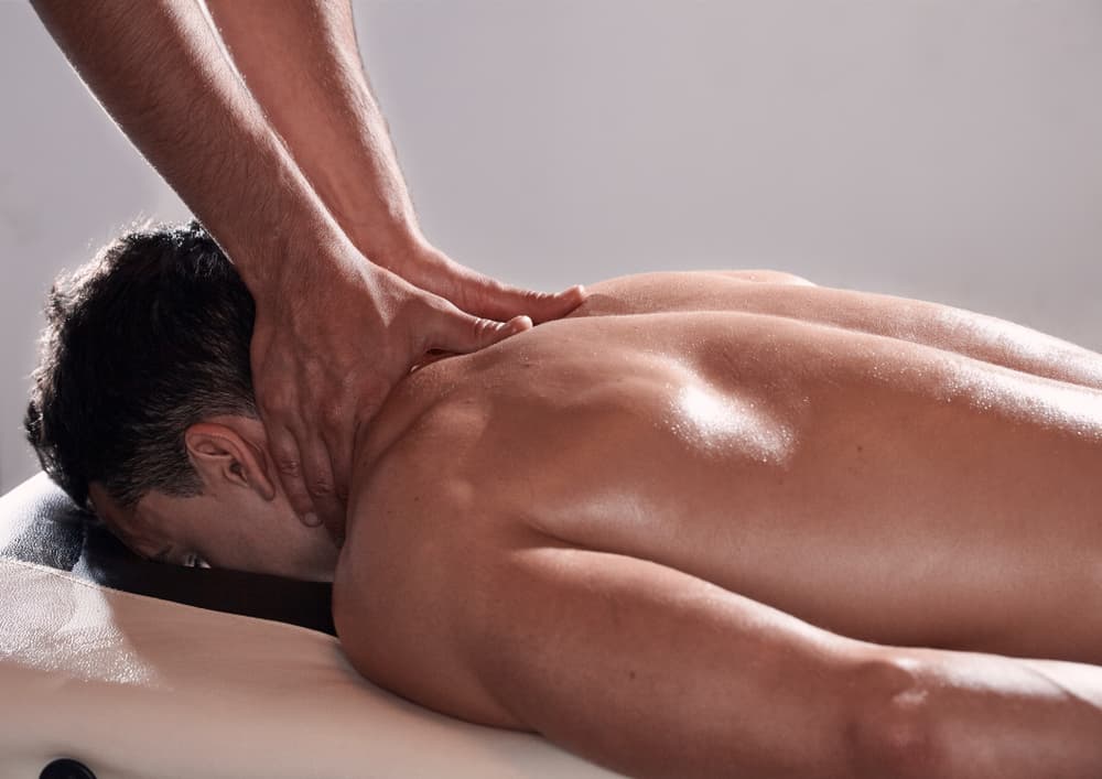 Man lying face down receiving a full body massage with oil, highlighting neck and upper back, in a calm and private treatment setting. Ideal for promoting combined massage and foot care treatment.