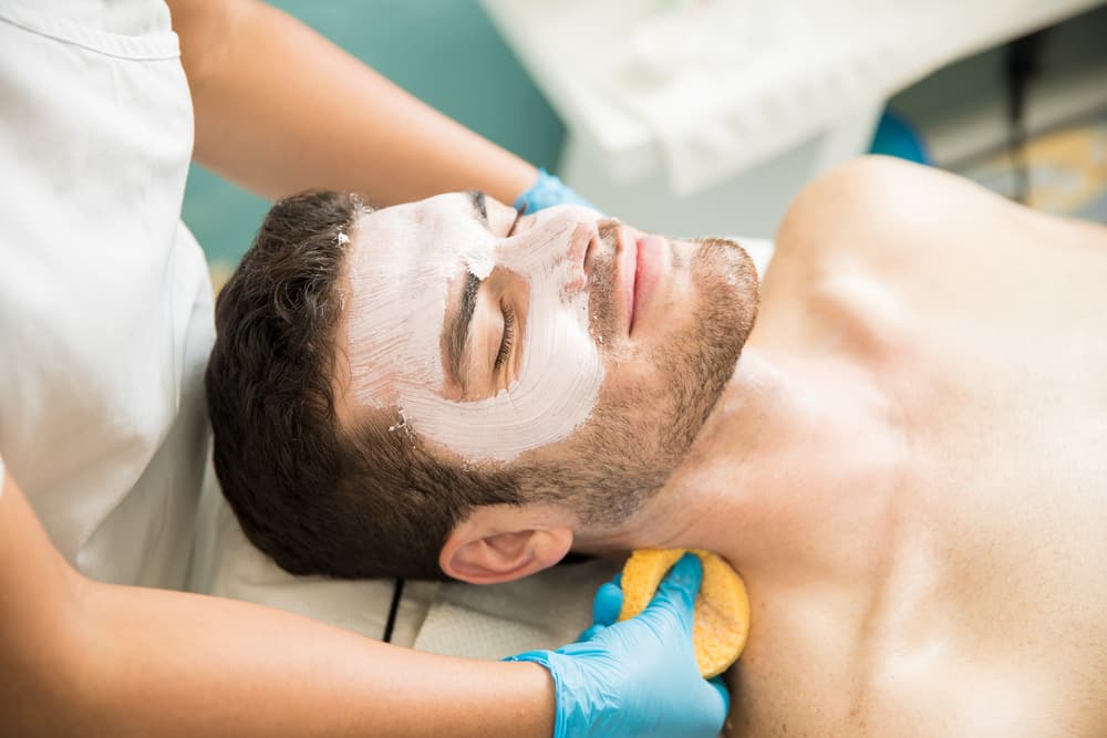 Man enjoying a relaxing facial treatment with a white face mask and sponge cleansing, delivered by a therapist in gloves. Ideal for promoting deluxe facials combined with full body massage.        Ask ChatGPT
