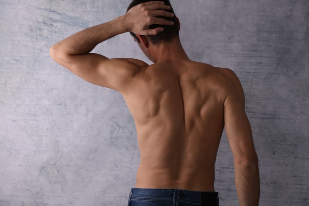 Therapist applying waxing strip to upper back of a male client lying face down in a treatment room.